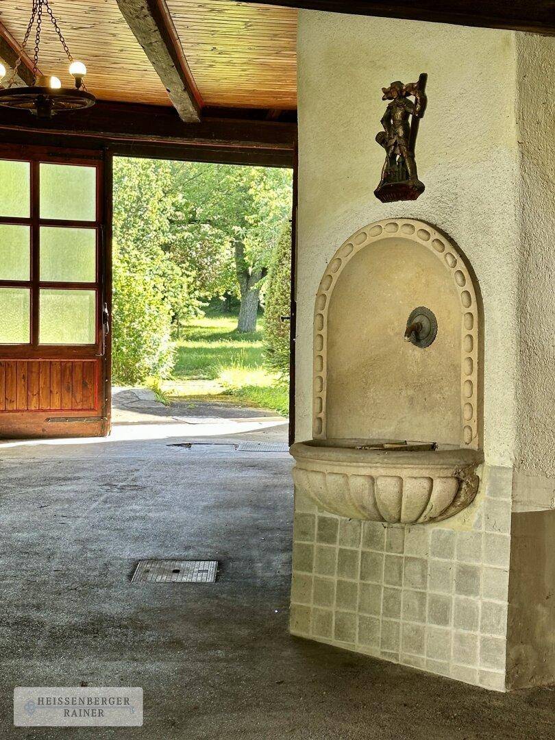 Überdachter Außenbereich mit einem dekorativen Steinbrunnen und Blick ins Grüne.