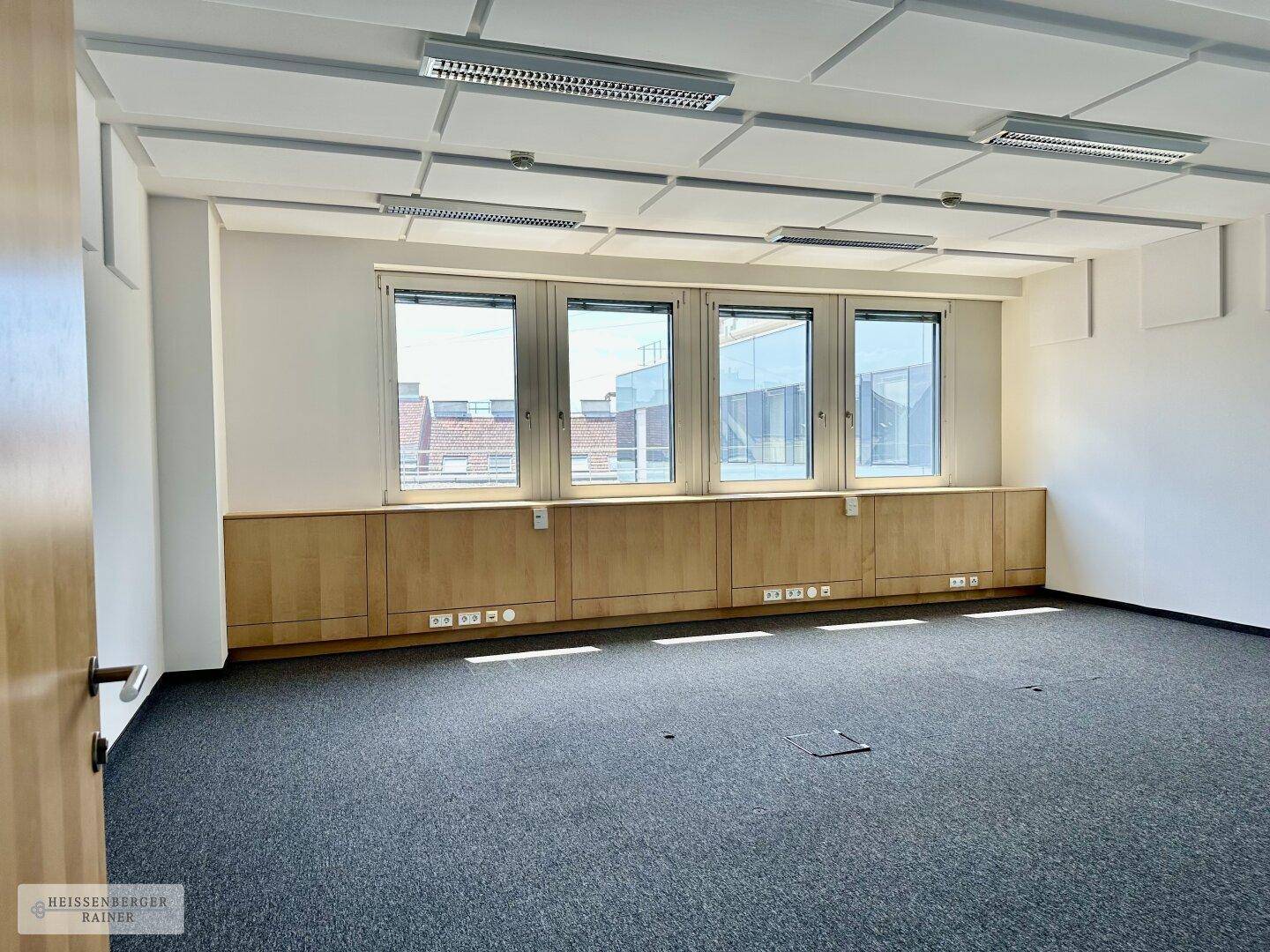 Heller Büroraum mit Akustikdeckenplatten, Teppichboden und Fenstern.