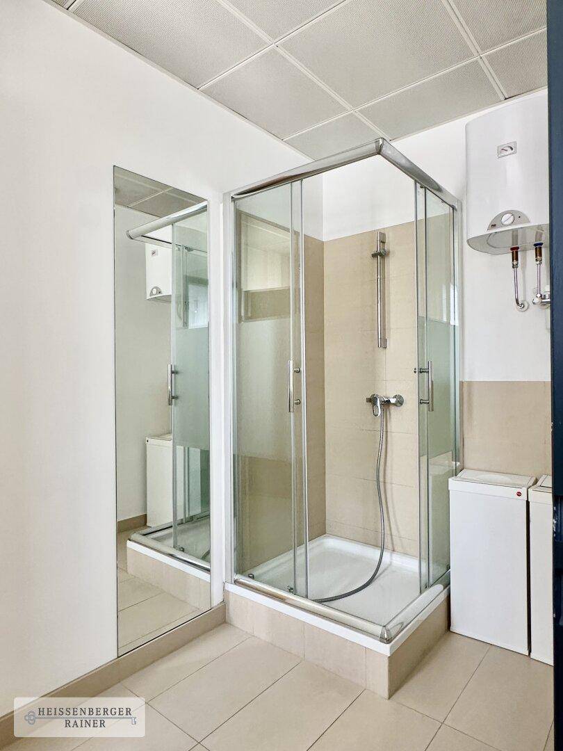 Badezimmer mit Duschkabine, beigen Fliesen und einem Warmwasserboiler.