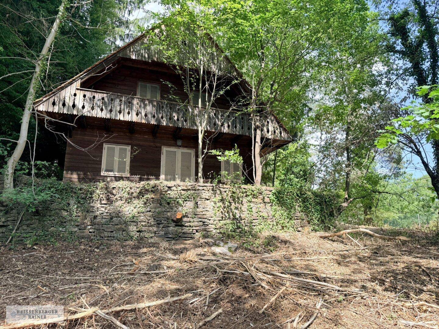 Rustikales Holzhaus mit verziertem Balkongeländer auf Steinfundament, umgeben von Bäumen und Wildwuchs.