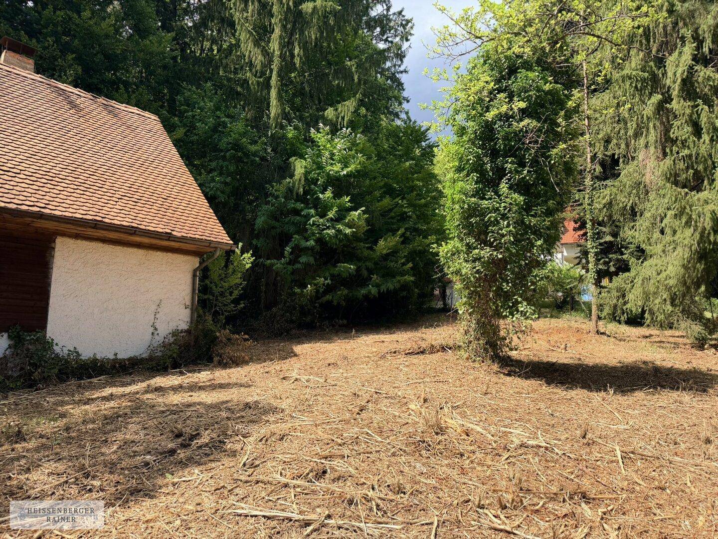 Teilansicht eines Hauses mit weißer Fassade und rotem Dach, umgeben von einem gerodeten Grundstück und Bäumen.