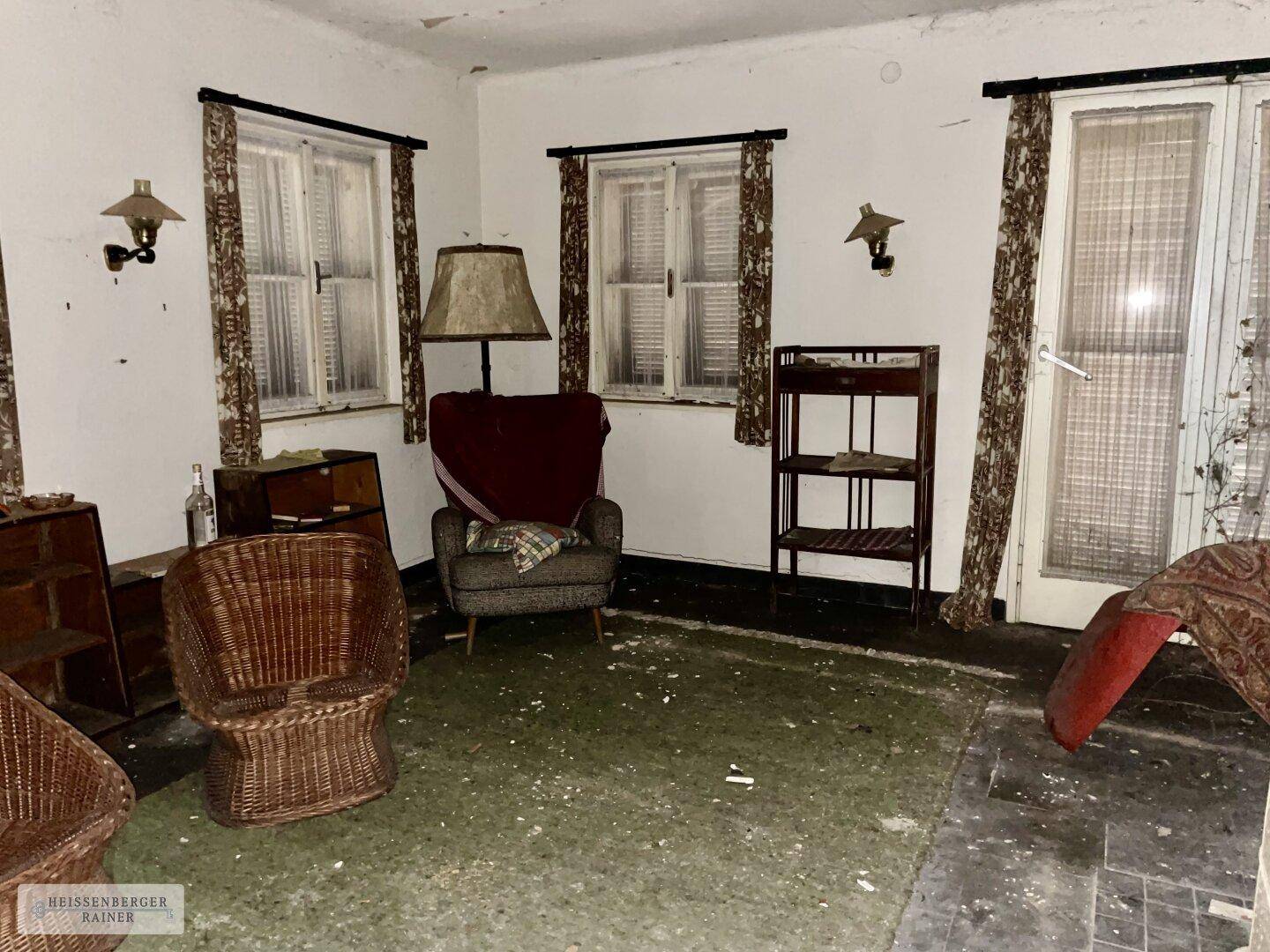 Vernachlässigtes Wohnzimmer mit alten Möbeln, zwei Fenstern mit Fensterläden und einer Tür.