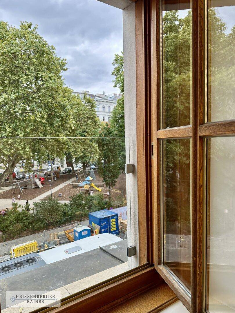 Blick aus dem Fenster auf einen belebten Platz mit Spielplatz und Bäumen.