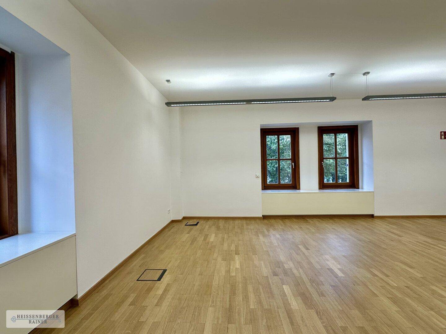Leerer Büroraum mit Holzboden und zwei Fenstern, bietet viel Tageslicht und eine angenehme Atmosphäre.