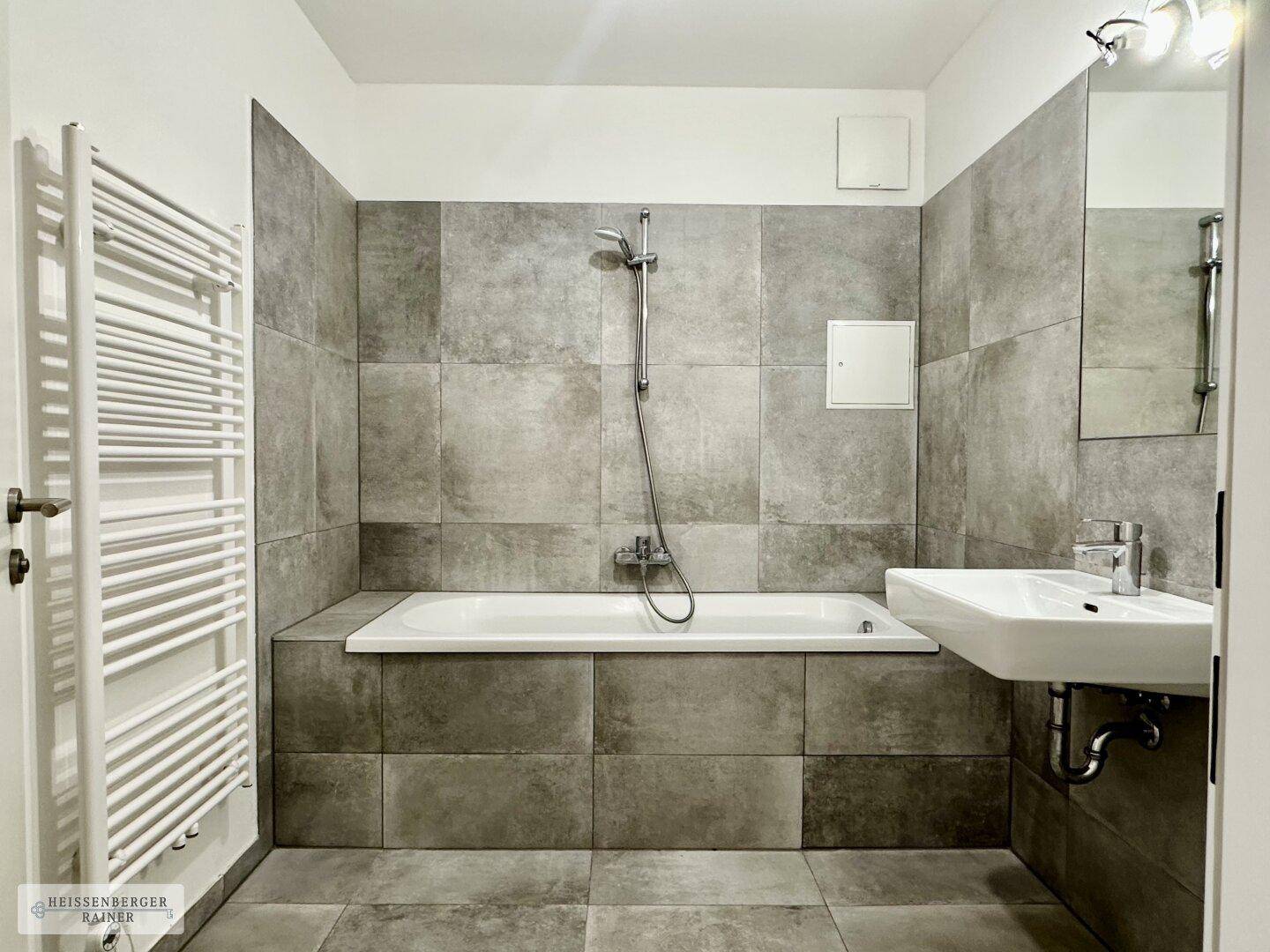 Modernes Badezimmer mit grauen Fliesen, Badewanne mit Duschfunktion und Handtuchheizkörper.