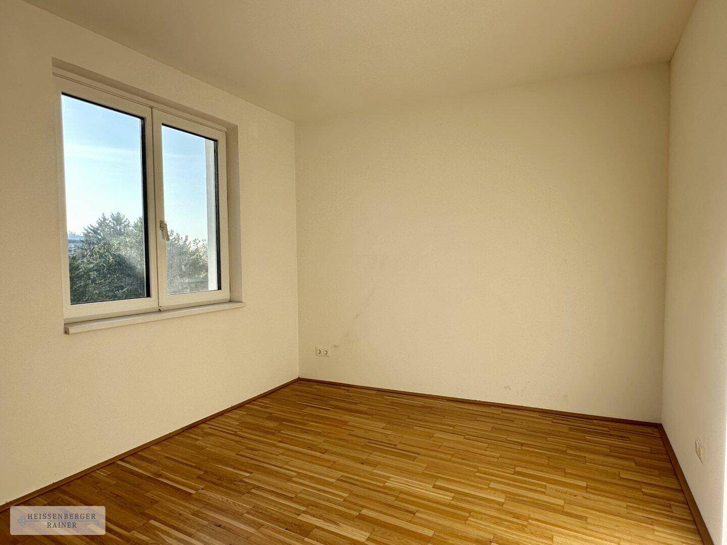 Geräumiges Zimmer mit Parkettboden und großem Fenster mit Blick auf Bäume und Natur.