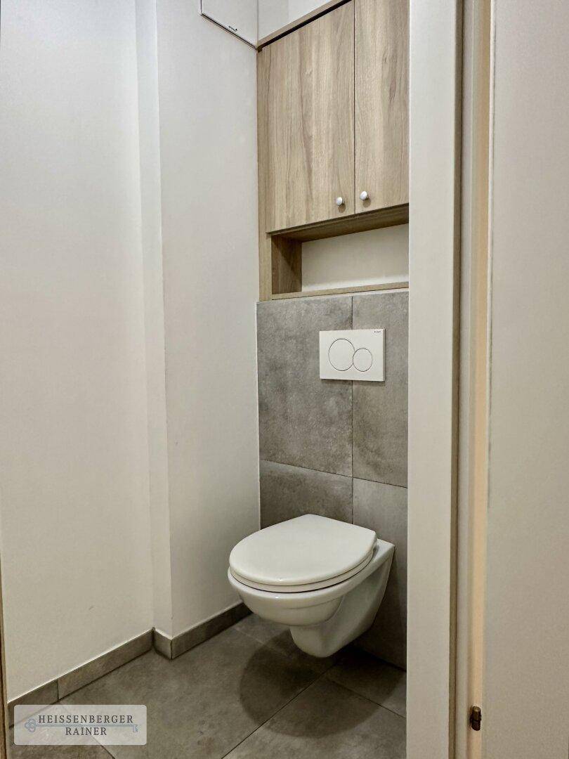 Modernes Gäste-WC mit wandhängender Toilette, grauen Fliesen und praktischem Wandschrank.