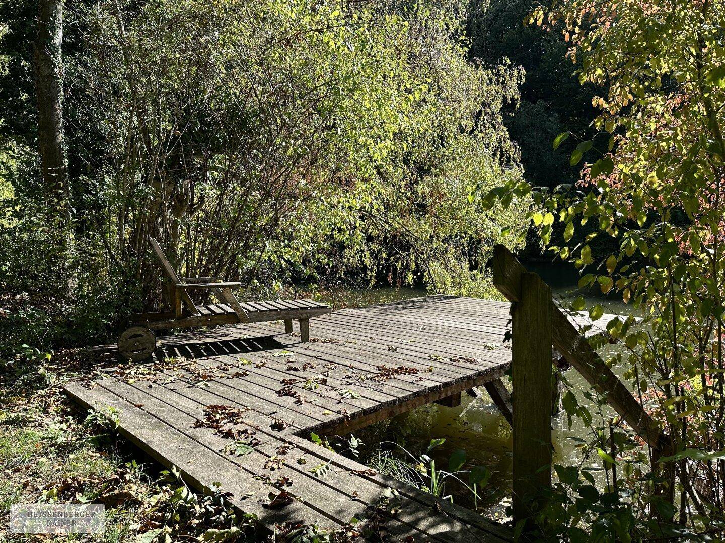Rustikale Holzterrasse mit Liegestuhl direkt am Wasser, umgeben von üppiger Vegetation.