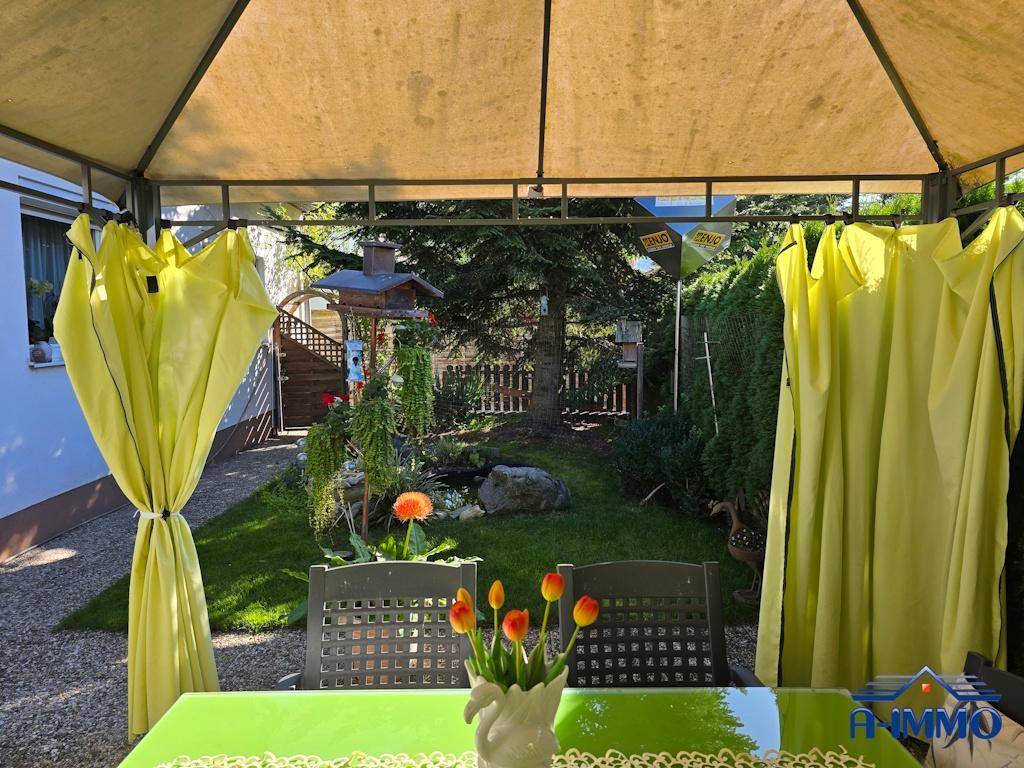 Blick aus dem Pavillon in den sonnigen Garten mit Blumen und Vogelhäuschen.