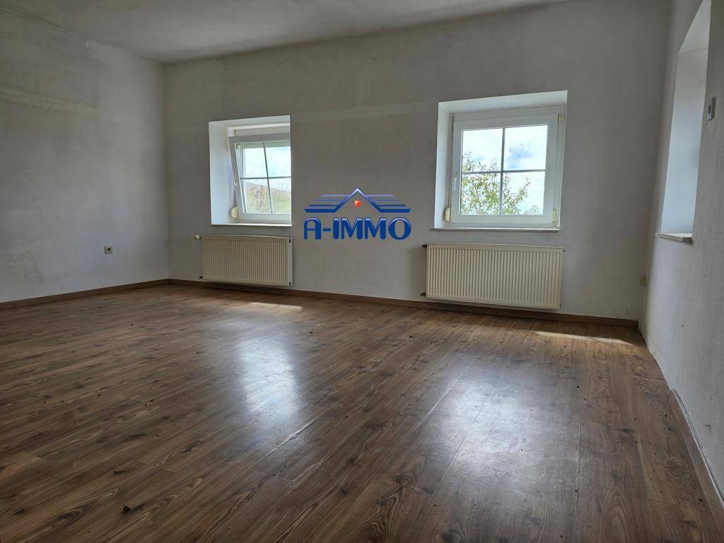 Leeres Wohnzimmer mit Laminatboden, weißen Wänden und zwei Fenstern mit Heizkörpern.