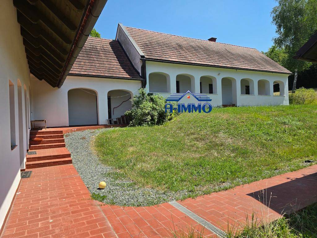 Rückseite des Hauses mit überdachter Arkade, gepflastertem Weg und grünem Hang.