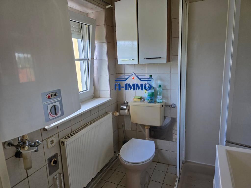 Badezimmer mit beigefarbenen Fliesen, Toilette, Duschkabine, Fenster und Warmwasserbereiter.