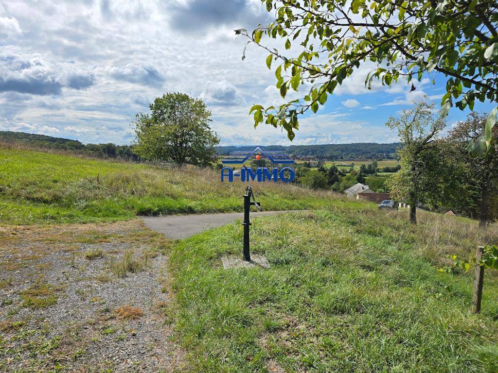 Ländliche Umgebung mit Kiesweg, Wasserpumpe und weitem Blick über ein Tal mit Häusern.