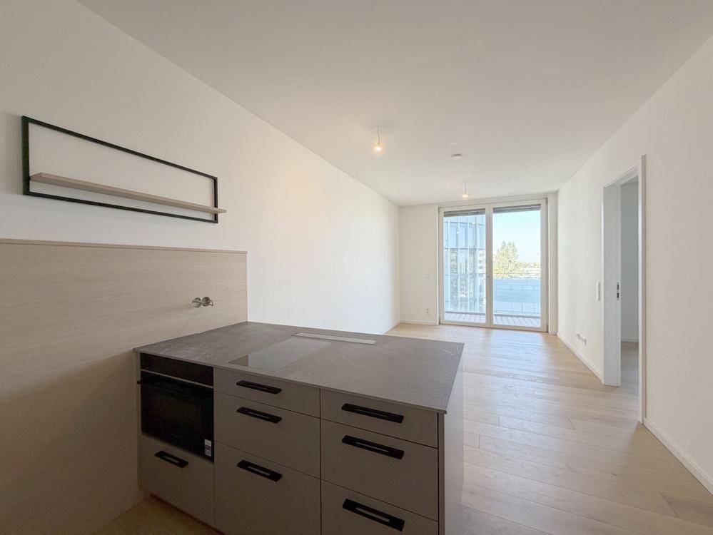 Offener Wohnbereich mit heller Holzboden, weißen Wänden und Zugang zu einem Balkon durch große Glasschiebetüren.