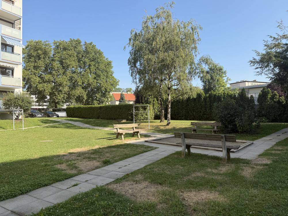 Grüne Gartenanlage mit Bäumen, Hecken und Holzbänken, ideal zum Entspannen im Freien.