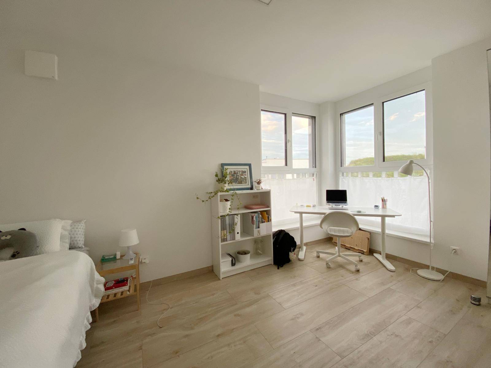 Helles Schlafzimmer mit Bett, Schreibtisch, Bücherregal und großen Fenstern für viel Tageslicht.