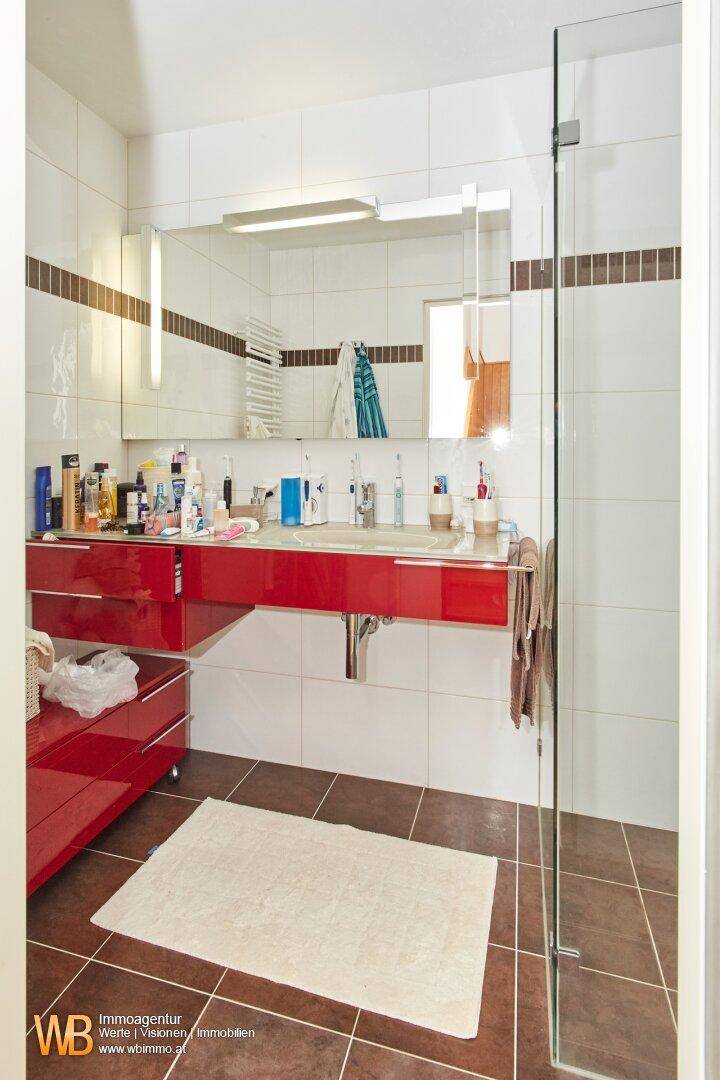 Badezimmer mit rotem Waschtisch, großem Spiegel, beheiztem Handtuchhalter und Duschbereich.