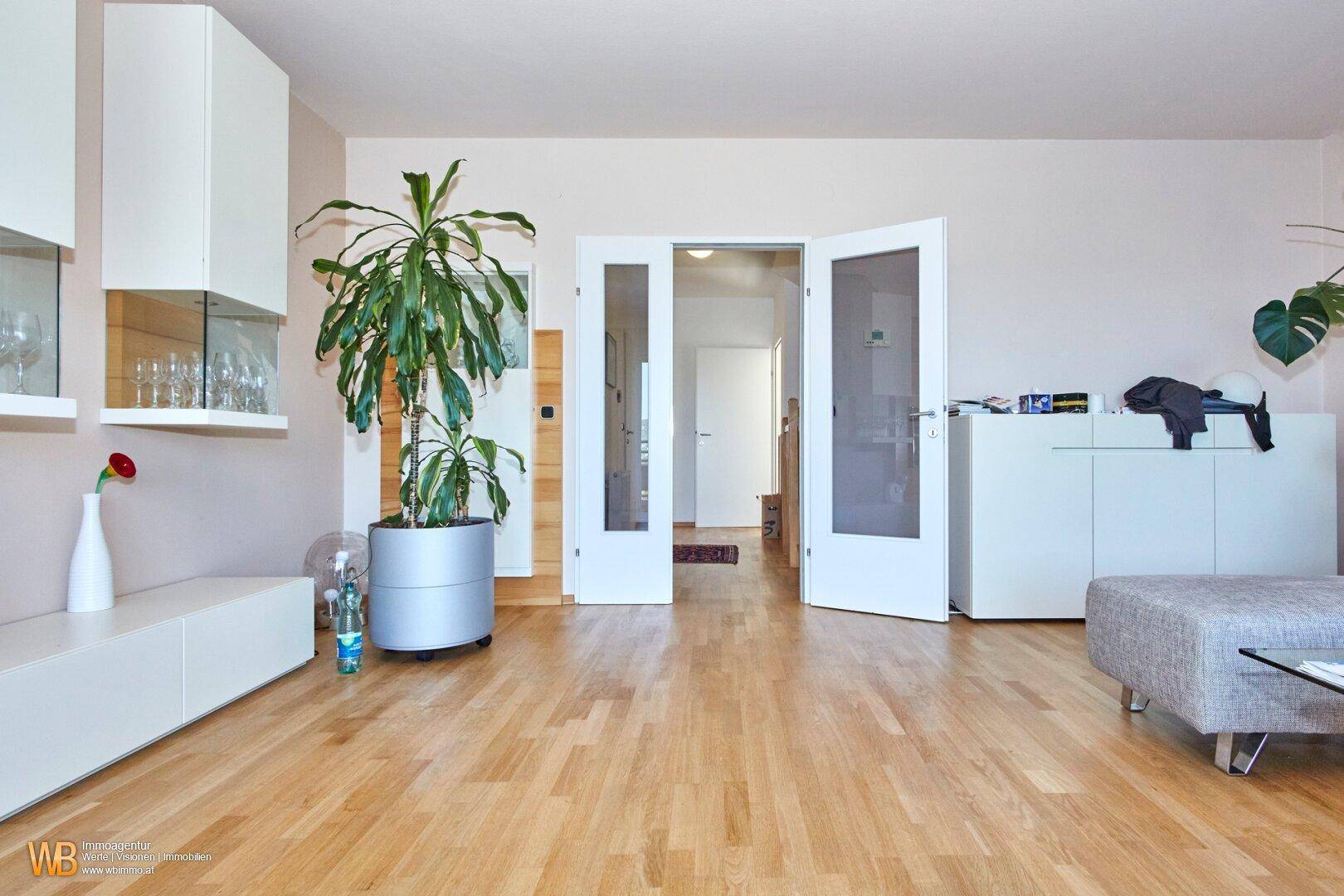 Geräumiges Wohnzimmer mit hellem Holzboden, Zimmerpflanze und Glastüren zu einem weiteren Raum.