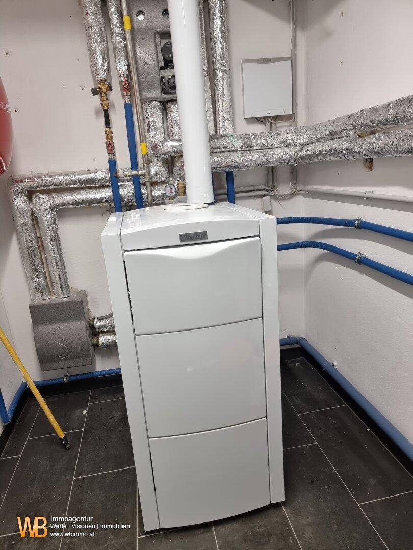 Heizungsanlage in einem Technikraum mit isolierten Rohren und dunklen Bodenfliesen, gut zugänglich.