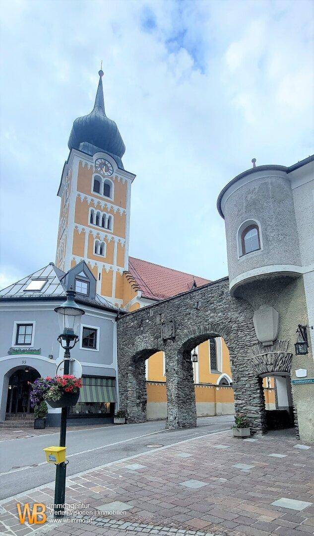 Historische Gebäudeansicht mit Kirchturm, Steinbogen und gepflastertem Weg.