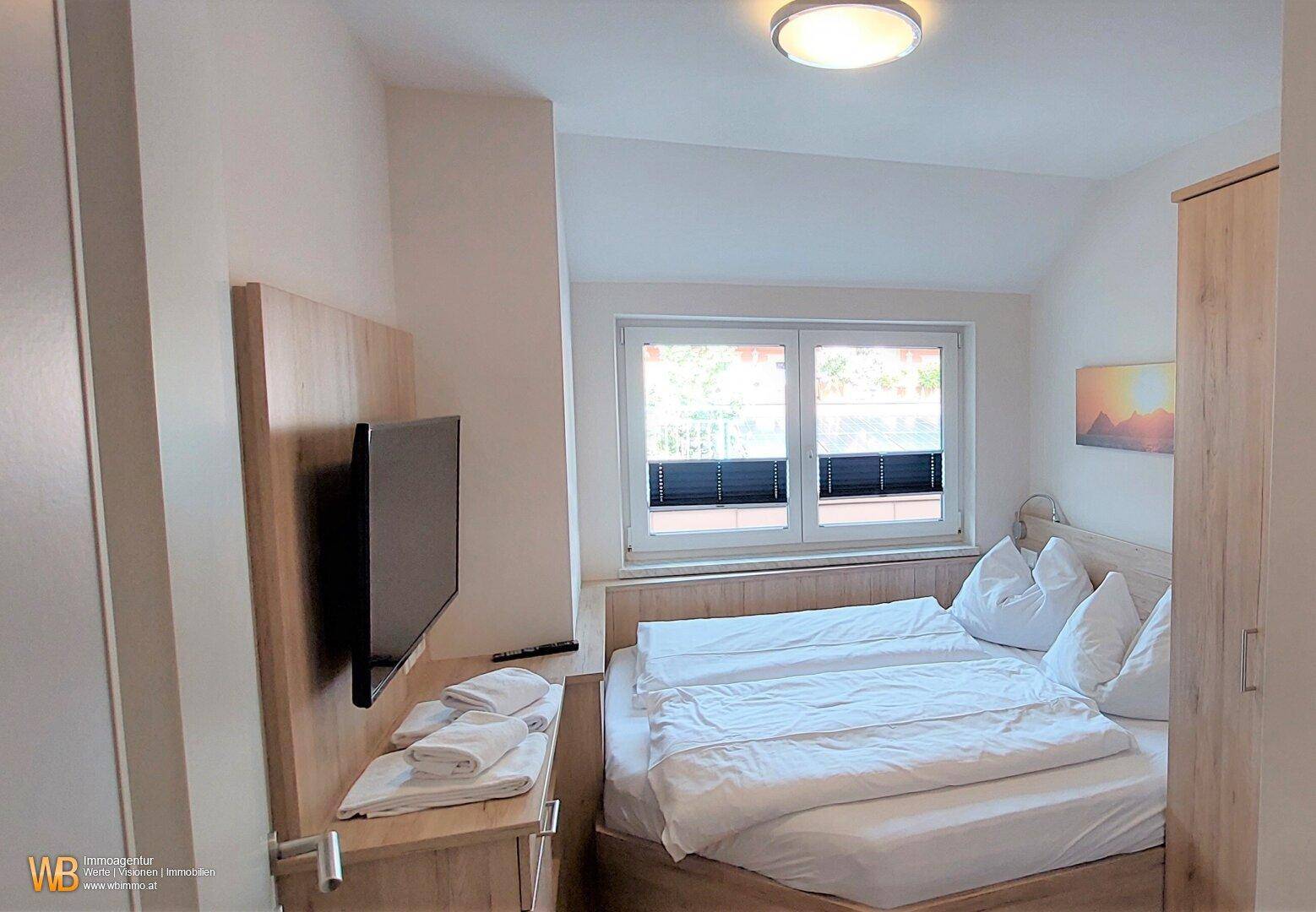 Helles Schlafzimmer mit Doppelbett, Fernseher und Fenster mit Jalousien.
