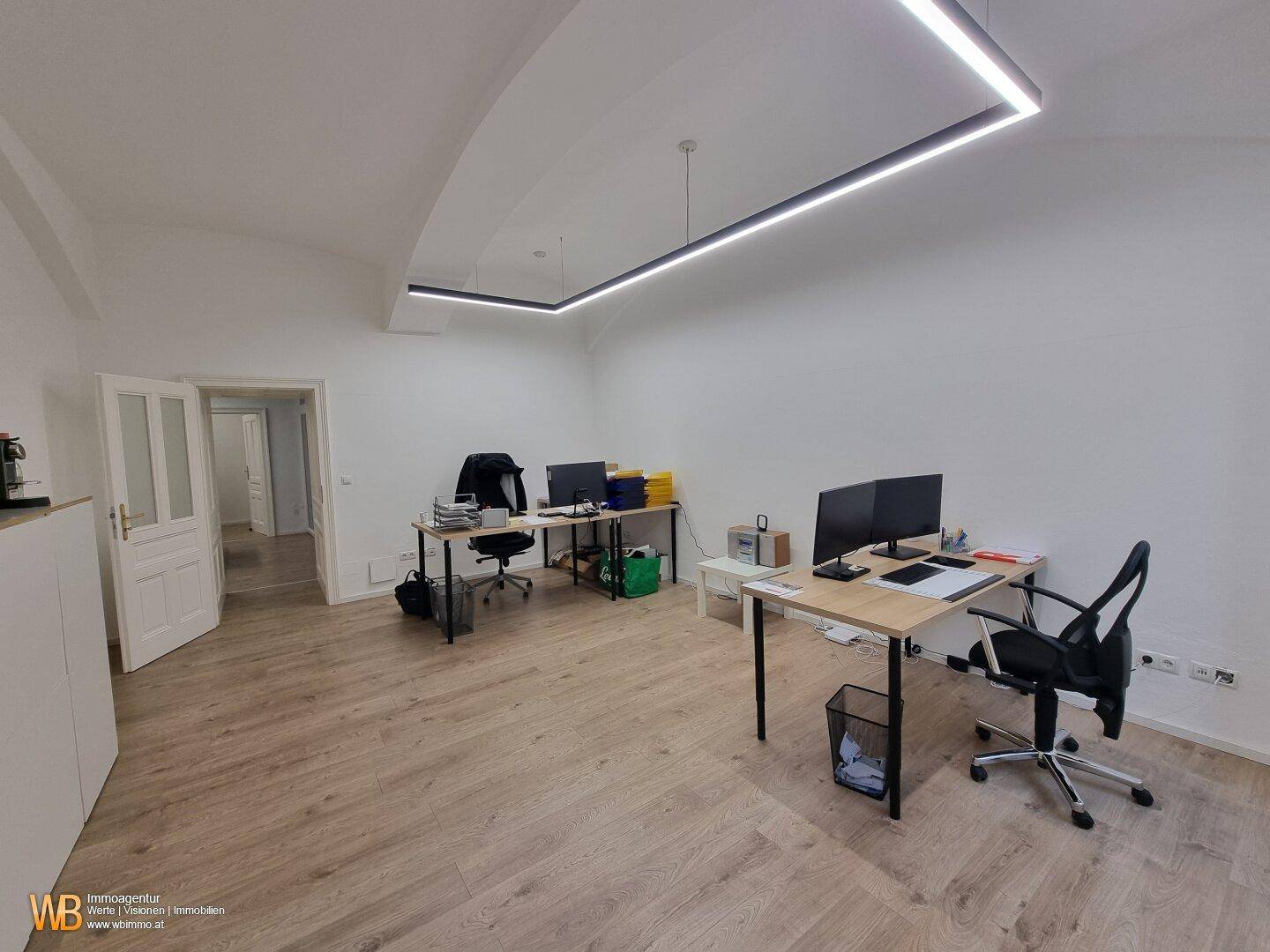 Offener Bürobereich mit mehreren Arbeitsplätzen, Holzboden und Zugang zu weiteren Räumen.