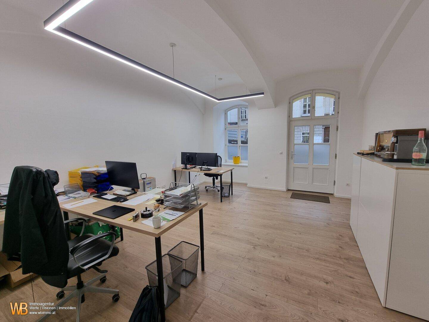 Heller Büroraum mit Holzboden, weißen Wänden und einem Eingangsbereich zur Straße.