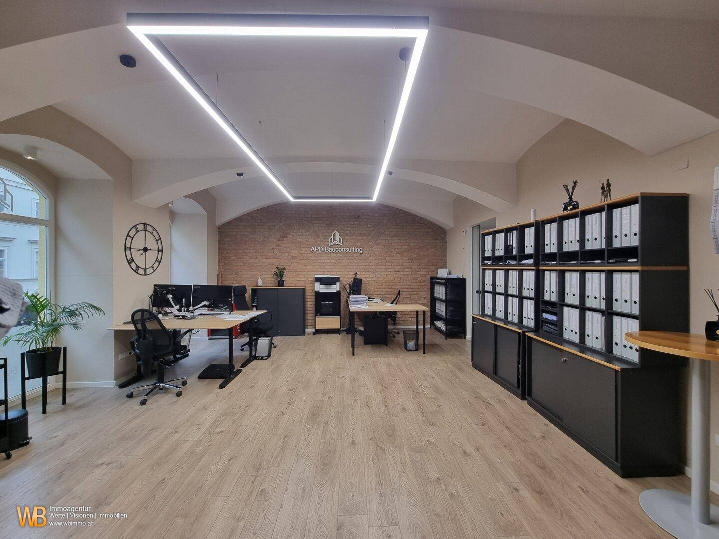 Weitläufiges Büro mit mehreren Arbeitsplätzen, Holzboden und einer markanten Ziegelwand.