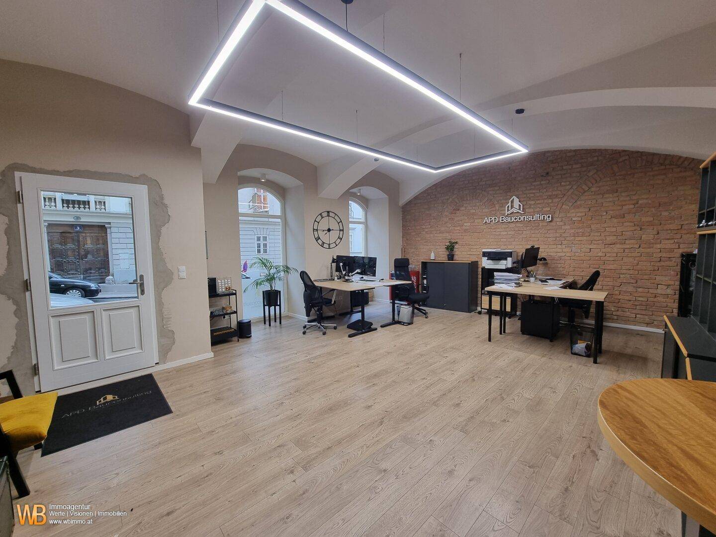 Helles Büro mit Holzboden, hohen Fenstern und einer Ziegelwand, die zur Straße hin ausgerichtet ist.