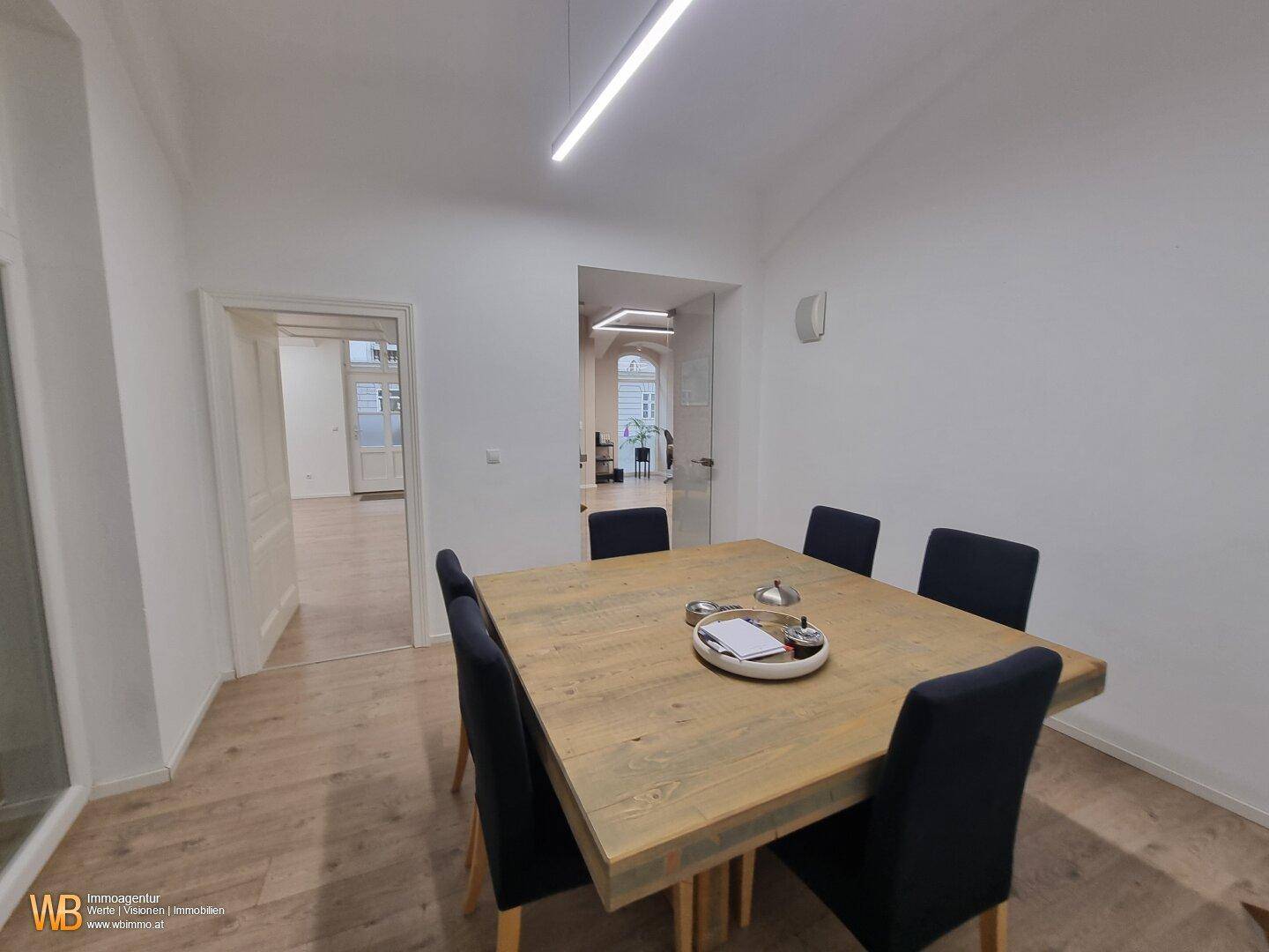 Besprechungsraum mit großem Holztisch und dunklen Stühlen, verbunden mit weiteren Büroräumen.