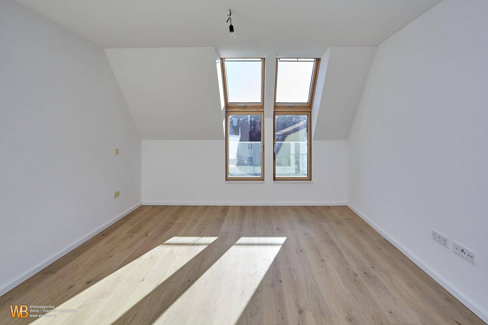 Helles Zimmer im Dachgeschoss mit schrägen Wänden, Holzboden und zwei großen Dachfenstern.