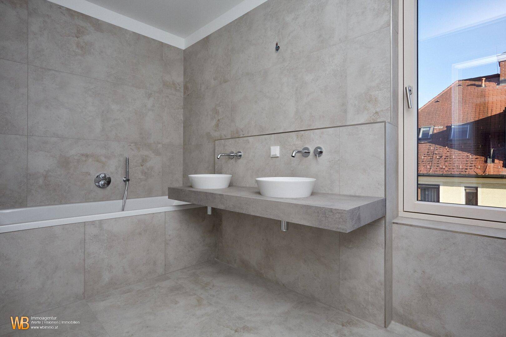 Modernes Badezimmer mit grauen Fliesen, Einbaubadewanne und schwebendem Doppelwaschtisch mit Fenster.