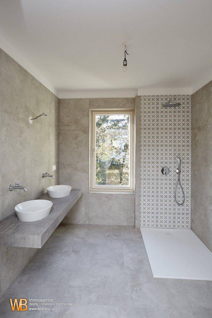 Modernes Badezimmer mit grauen Fliesen, begehbarer Dusche und schwebendem Doppelwaschtisch mit Fenster.
