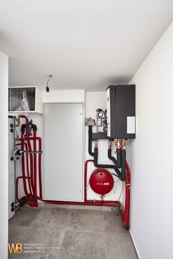 Technikraum mit Heizungsanlage, Warmwasserspeicher und sichtbaren Rohrleitungen auf Fliesenboden.