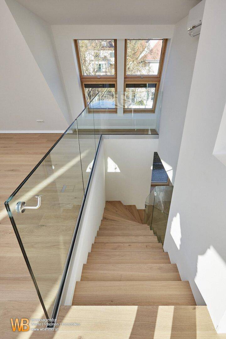 Moderne Holztreppe mit Glasgeländer und viel Tageslicht durch die darüberliegenden Dachfenster.