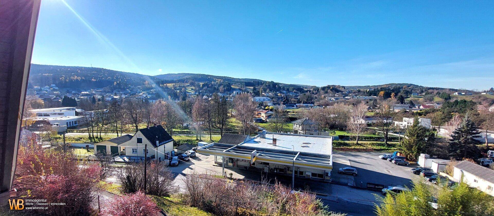 Weitläufiger Blick über die Siedlung mit einer Tankstelle und umliegenden bewaldeten Hügeln.