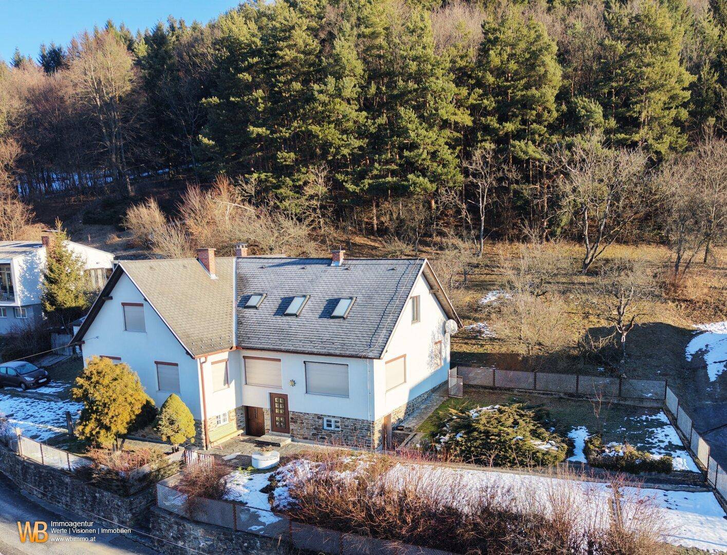Luftaufnahme eines Einfamilienhauses mit Satteldach und umliegendem Garten in bewaldeter Umgebung.