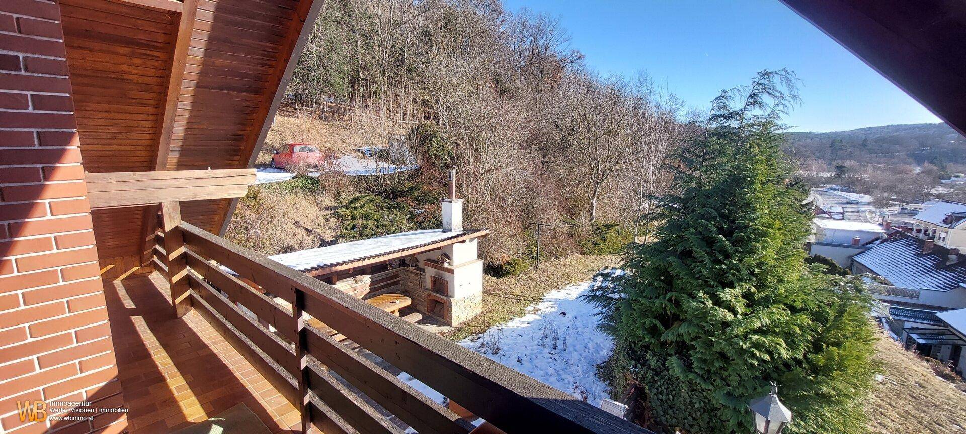 Balkon mit Ziegelwand und Holzgeländer, bietet einen Blick auf die bewaldete Hanglage und eine Grillstelle.