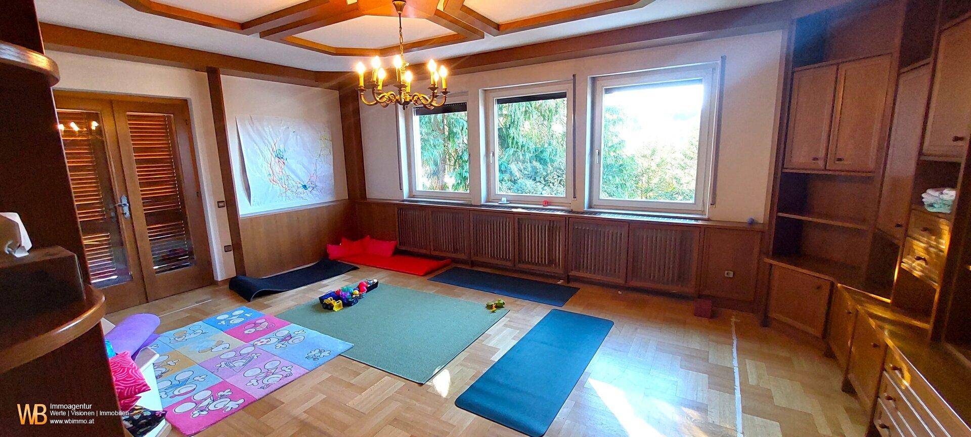 Geräumiges Zimmer mit Holzelementen, Parkettboden und großen Fenstern mit Blick ins Grüne.