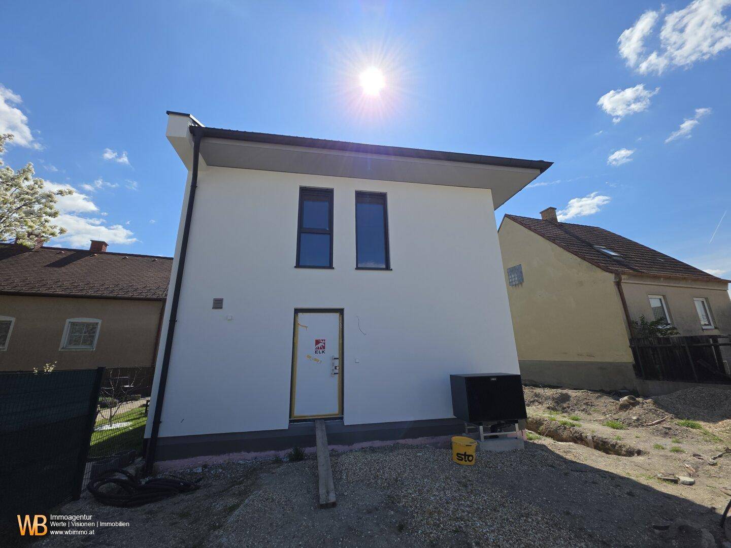Rückseite des Neubaus mit weißer Fassade, Eingangstür und externer Heizungsanlage.