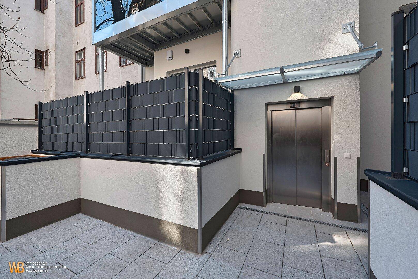 Außenbereich mit modernem Aufzug und gepflastertem Zugang, umgeben von einer Sichtschutzwand.