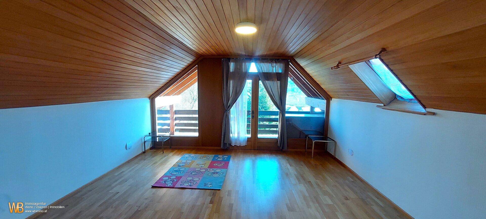 Helles Dachzimmer mit Holzverkleidung, Parkettboden und großen Fenstern, die viel Licht hereinlassen.