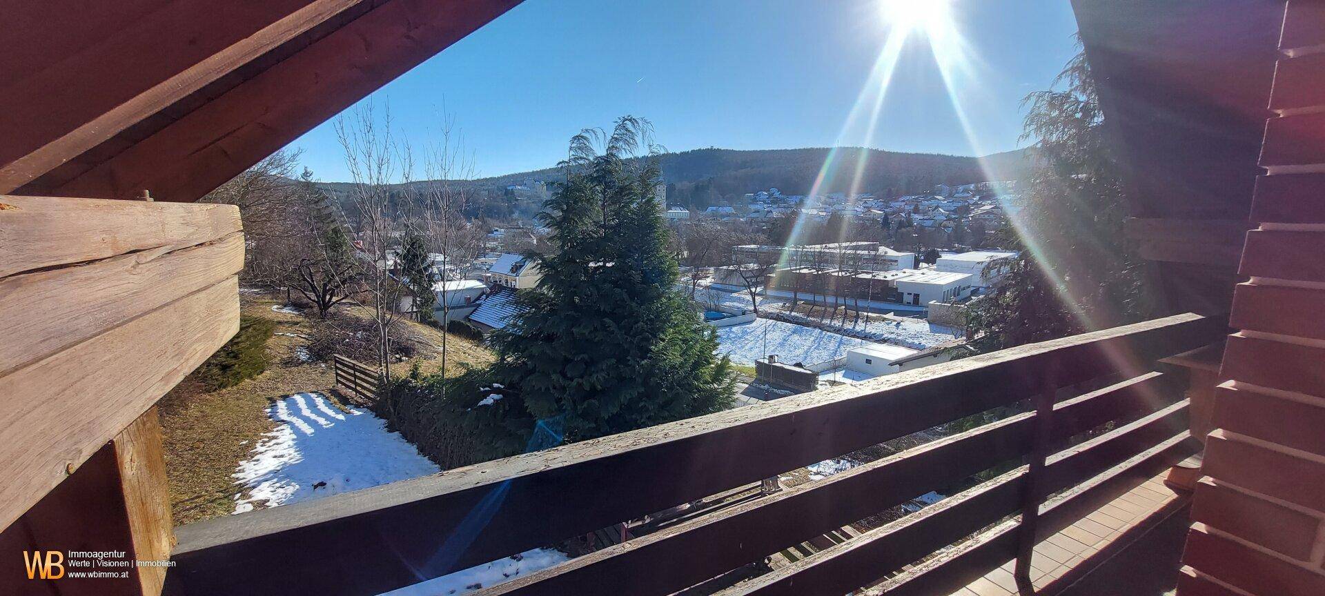Sonniger Ausblick von einem Holzbalcon auf eine verschneite Landschaft mit Bäumen und Häusern.