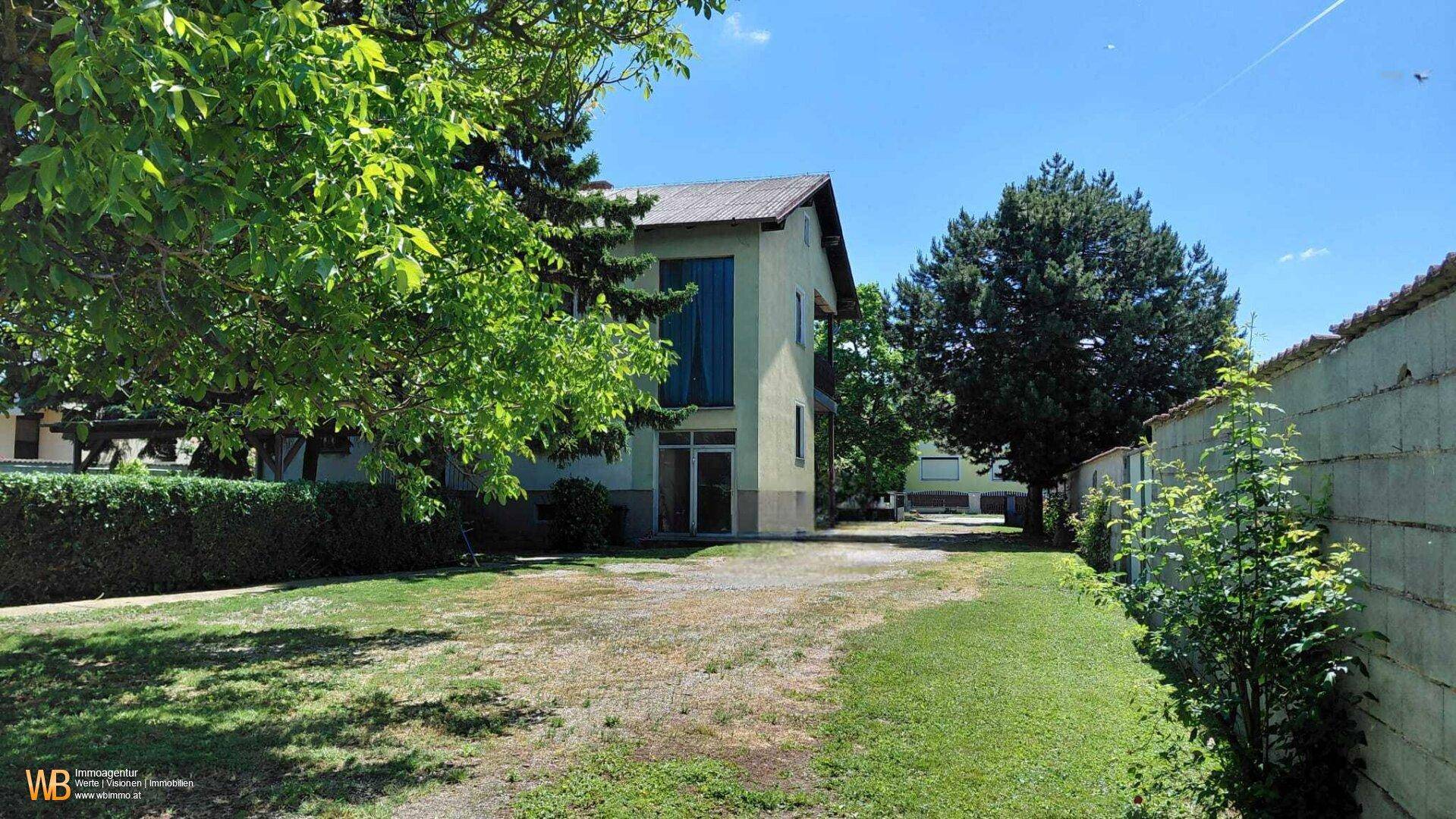 Seitenansicht des Hauses mit weitläufigem Grundstück, Bäumen und einer unbefestigten Zufahrt.