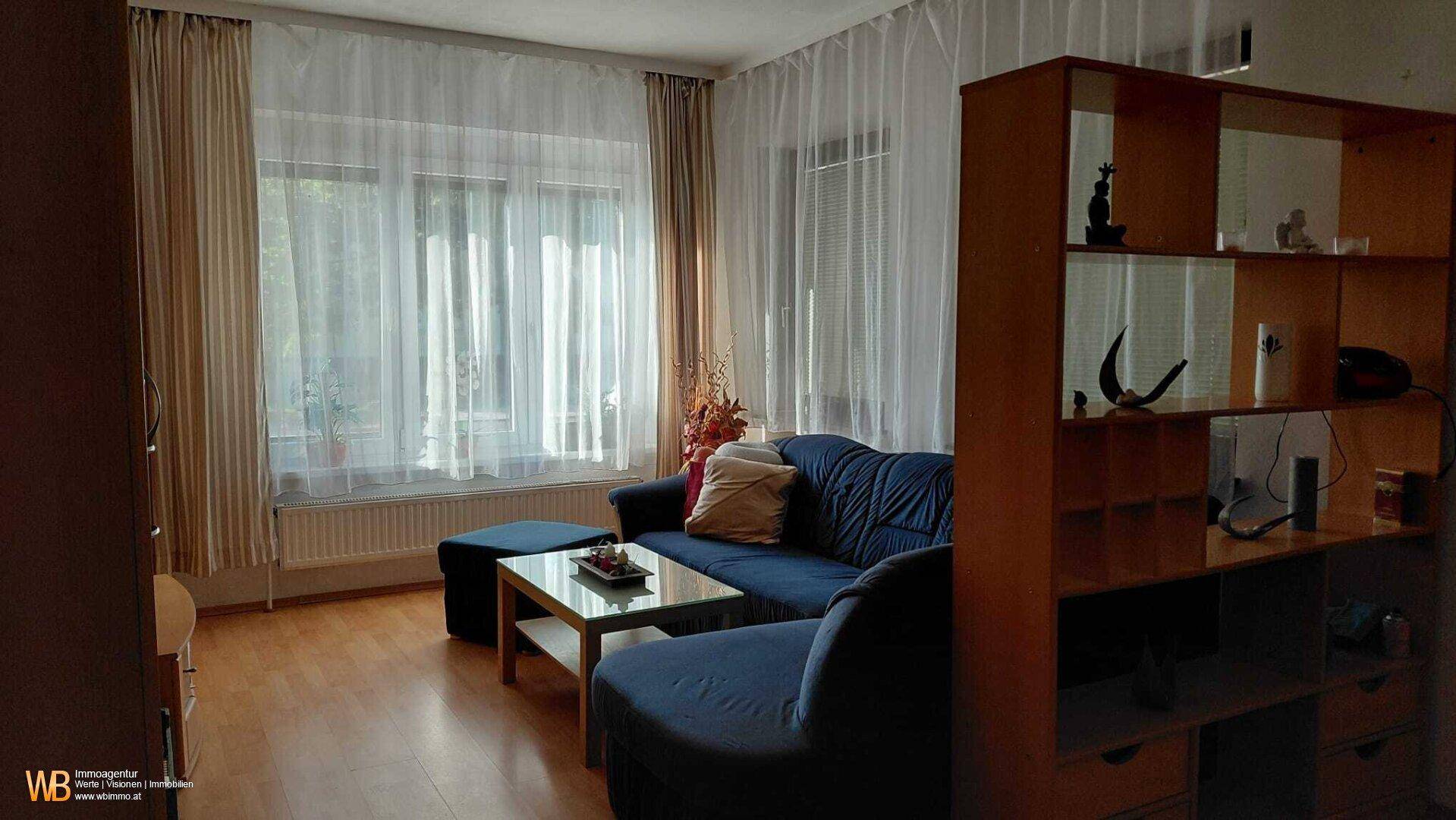 Gemütliches Wohnzimmer mit blauem Sofa, Couchtisch und großen Fenstern für viel Tageslicht.