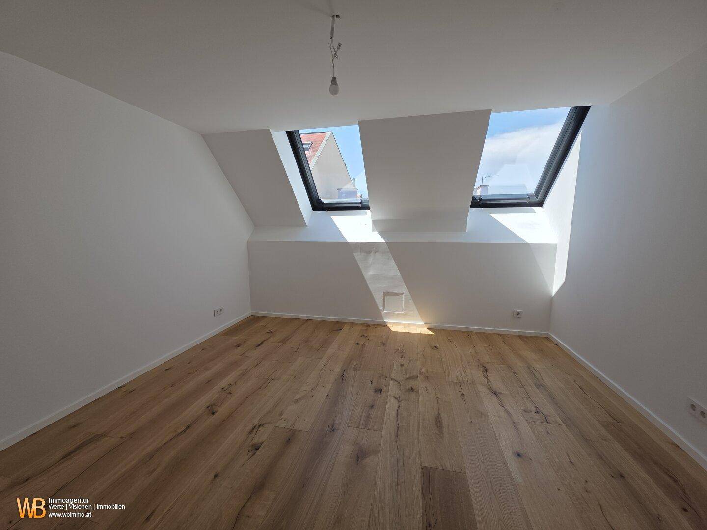 Helles Dachzimmer mit Holzboden und zwei großen Dachfenstern, die viel Tageslicht hereinlassen.