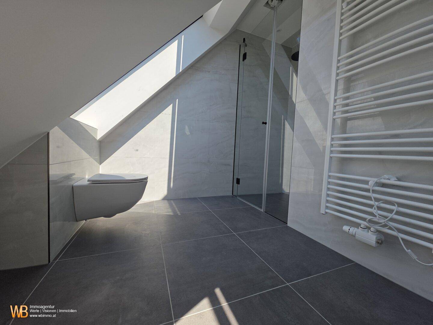 Duschbereich und wandhängendes WC im hellen Dachgeschossbad mit grauen Fliesen und Tageslicht.