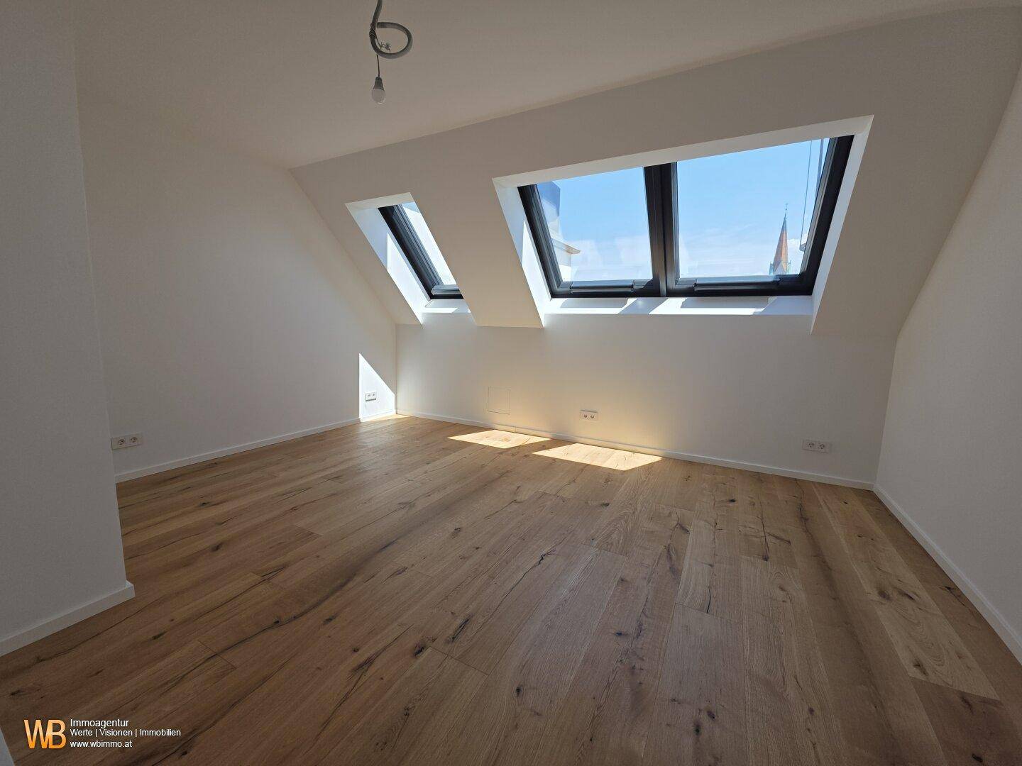 Helles Dachzimmer mit Holzboden und zwei großen Dachfenstern, die viel Tageslicht hereinlassen.