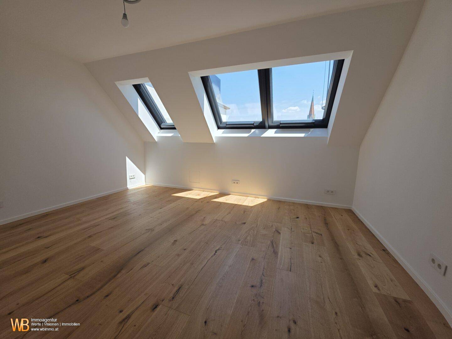Helles Dachzimmer mit Holzboden und zwei großen Dachfenstern, die viel Tageslicht hereinlassen.