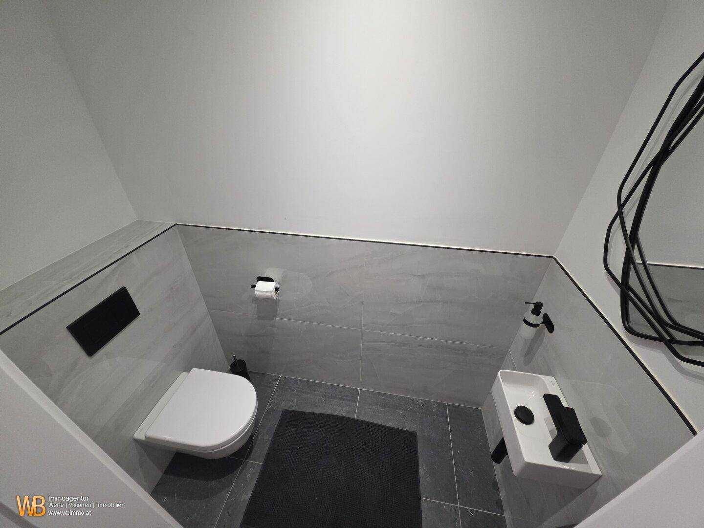 Modernes Gäste-WC mit grauen Fliesen, wandhängendem WC und kleinem Waschbecken unter der Dachschräge.